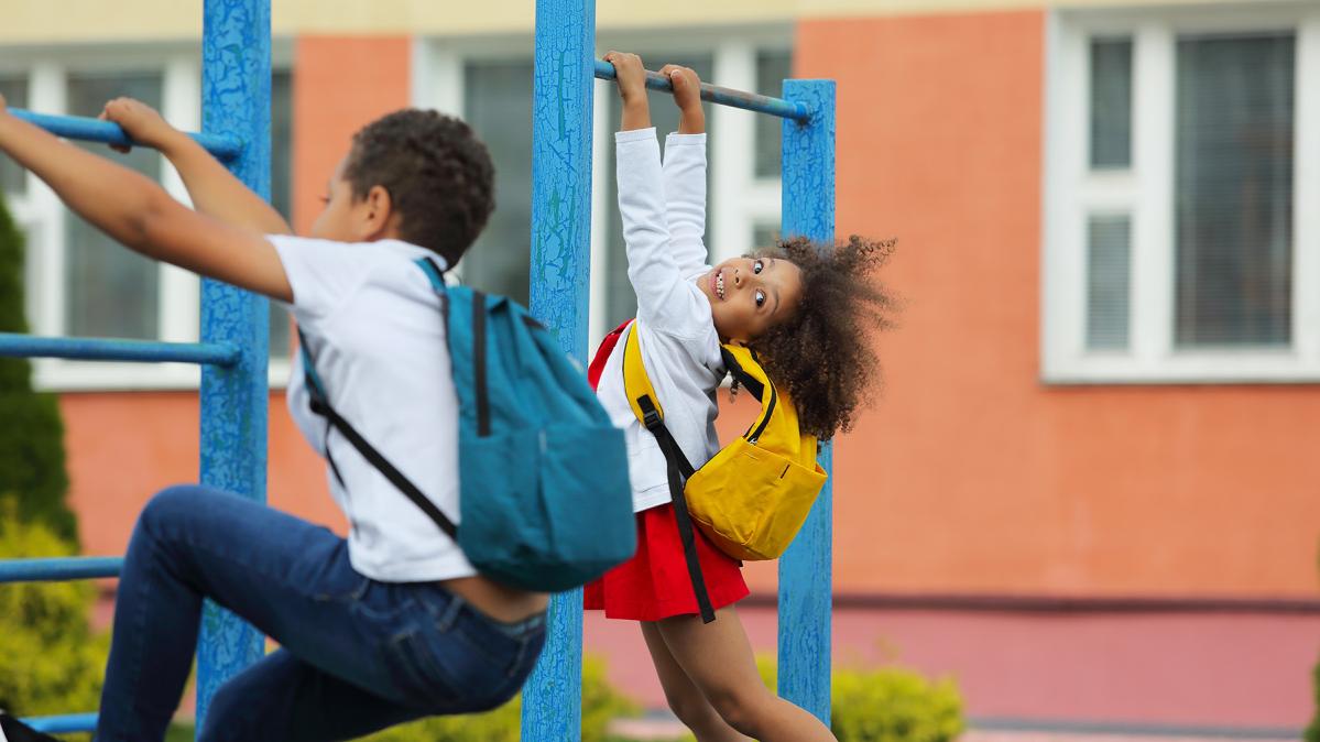 Two children wearing backpacks play on monkey bars