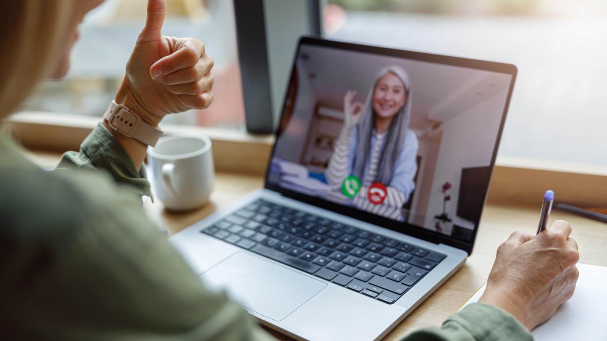 Cropped closeup of woman giving thumbs up to screen on Teams meeting on laptop while sitting in bay window of house.