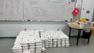 Stacks of white take-out boxes on a grey linoleum floor in front of a large, rectancular white board with lists written on it and a small table to the right on which sits a basket of coloured materials.