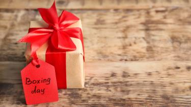Photo of smallish gift wrapped in brown paper with red ribbon and red label with boxing day written on it in black pen, with the gift sitting on a weathered, wooden plank background that could be a floor or a table top.