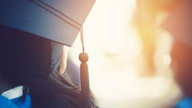 A photo looking close up at the back of the heads and shoulders of two students wearing blue gowns and black graduation caps with the sun beaming out between them. 