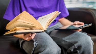 Literacy Child in purple shirt and grey jeans sitting holding an open book in one hand and tablet in the other.