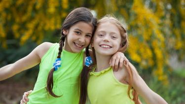 International Day of the Girl Child Two young girls wearing bright green and yellow sleeveless shirts and smiling broadly with arms around one anothers shoulders outdoors