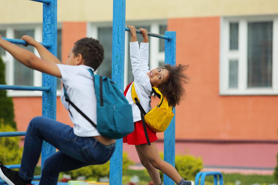 Two children wearing backpacks play on monkey bars